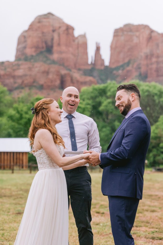 A man wearing a dark blue suit laughs during his Sedona wedding at Red Rock Crossing. He is holding hands with his bride who is smiling at him. Their officiant is behind them laughing and the Cathedral Rock is also behind them.