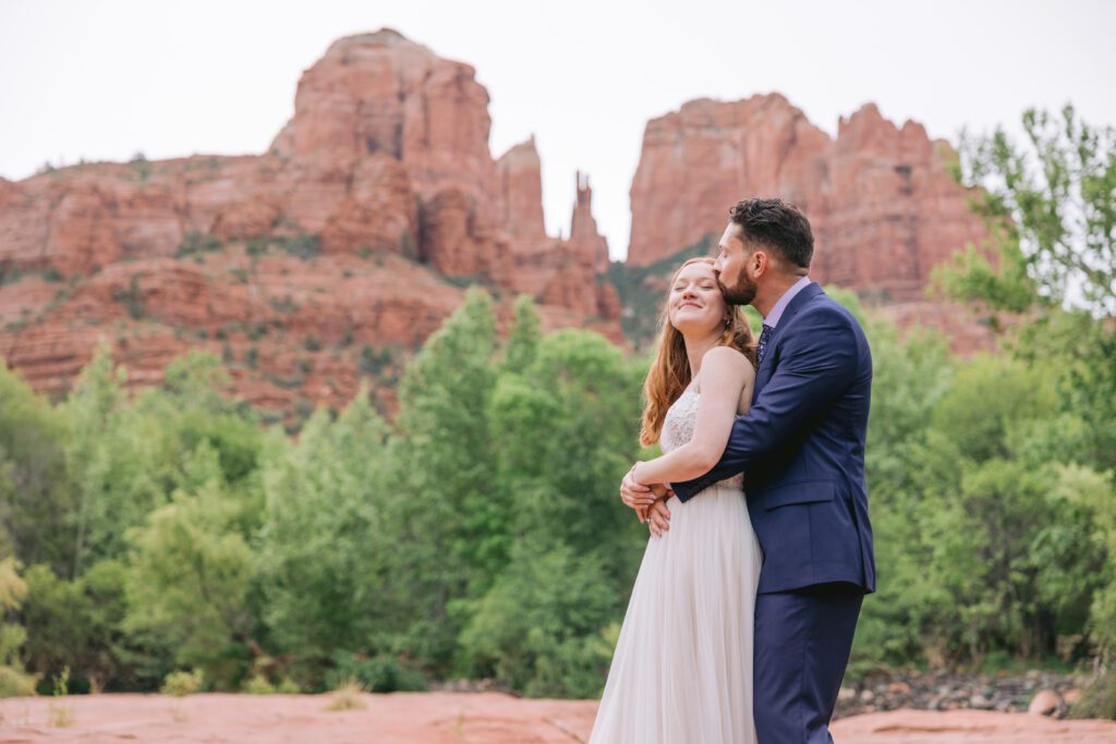 A bride with red hair and white wedding dress is embraced by a groom with dark hair wearing a dark blue suit. Green trees and Cathedral Rock are in the background behind them at their elopement at Red Rock Crossing.