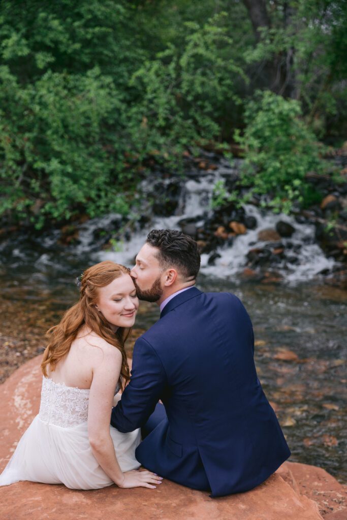 A man in a dark blue suit kisses the forehead of a woman with red hair wearing a wedding dress. A waterfall and water from Oak Creek are behind them.