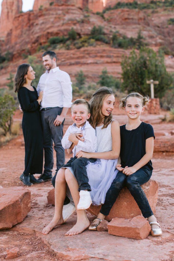 Three kids sit on a rock and smile while their parents are in the background for their family photos in Sedona.