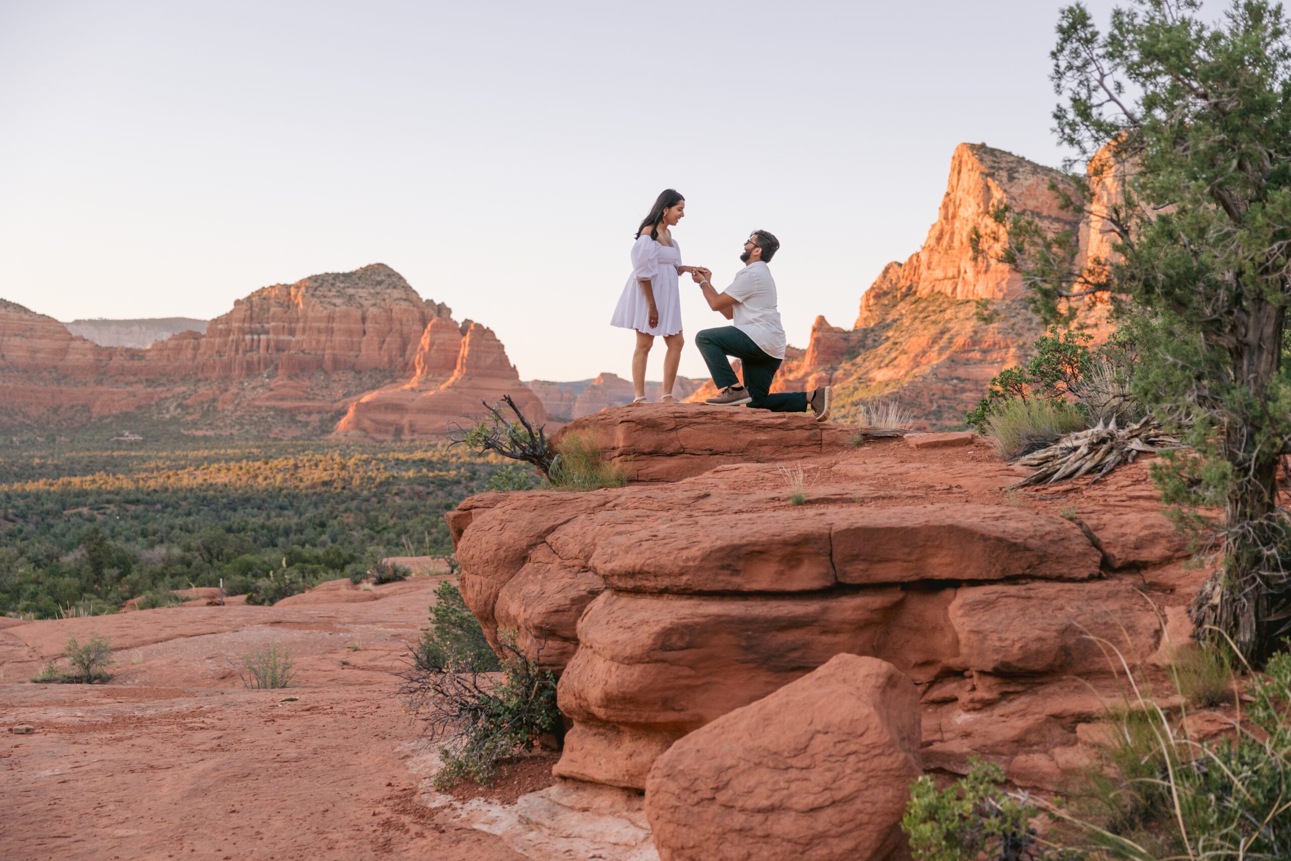 Surprise proposal in Sedona. A man wearing a white shirt is kneeled down on one knee as he proposes to a woman in a white dress with the red rocks behind them.