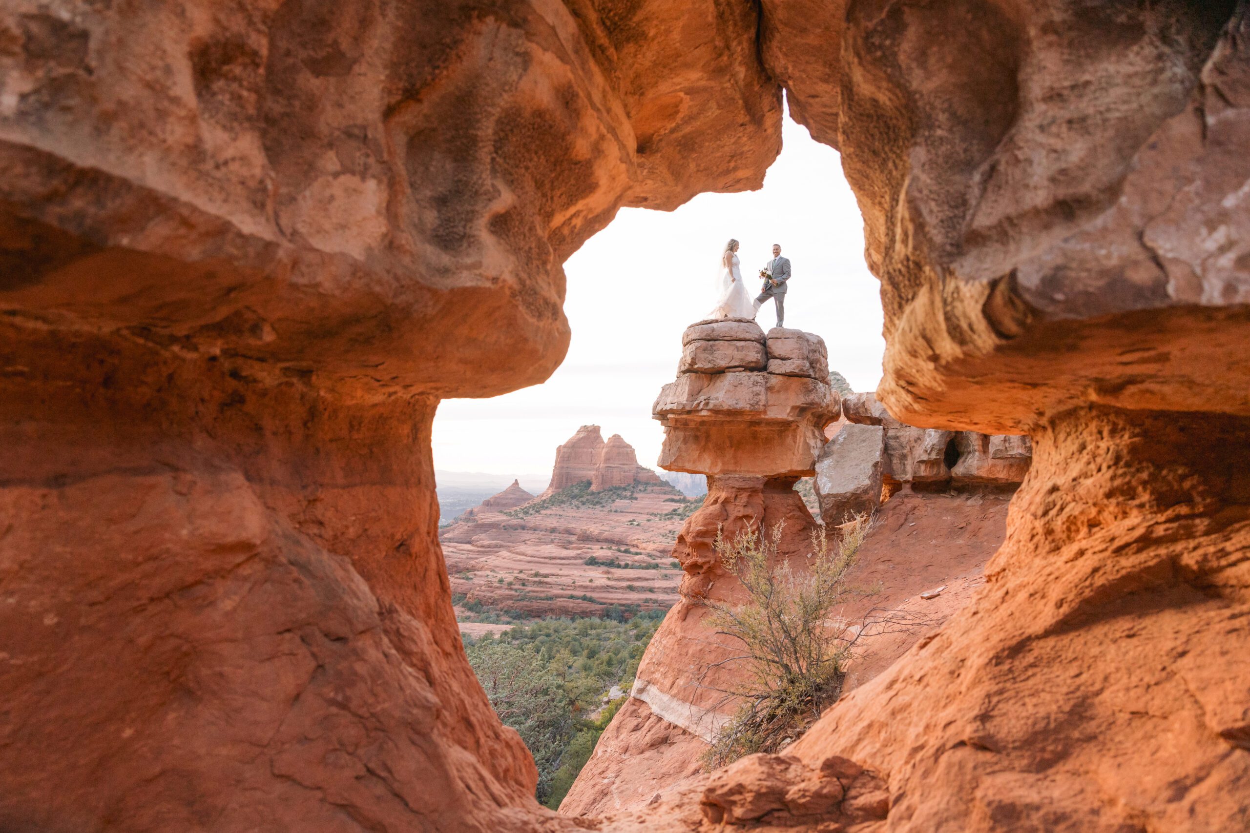 Sedona adventure elopement at Merry Go Round. A bride and groom stand on a red rock while the photographer photographs them through an opening in another set of red rocks.