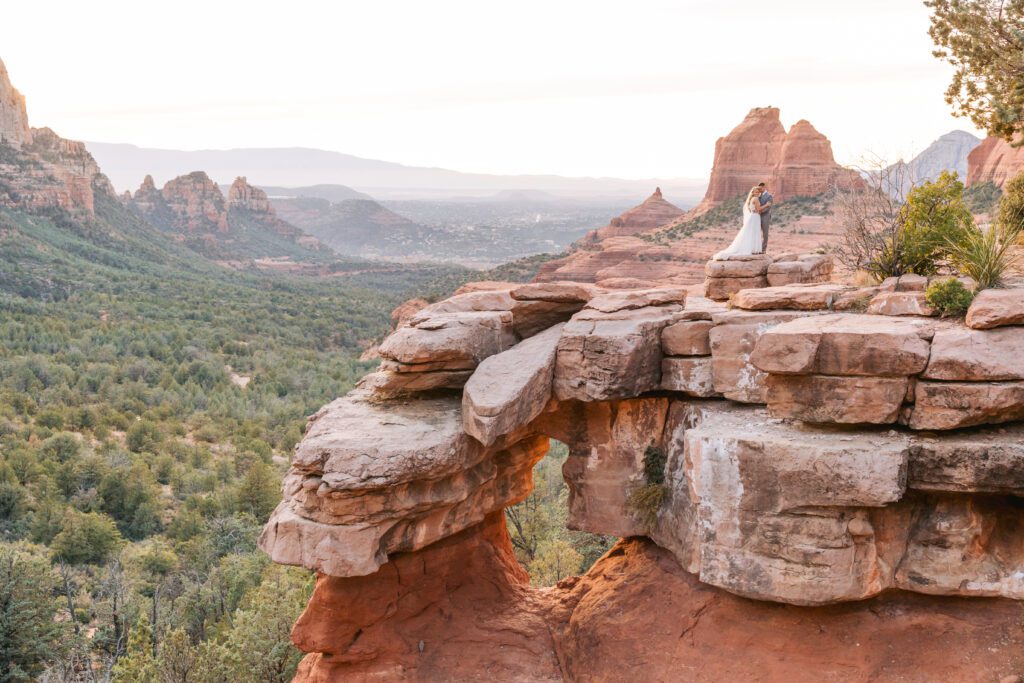 A couple stands on a ledge on Merry Go Round Rock in their wedding attire on their Sedona elopement day.