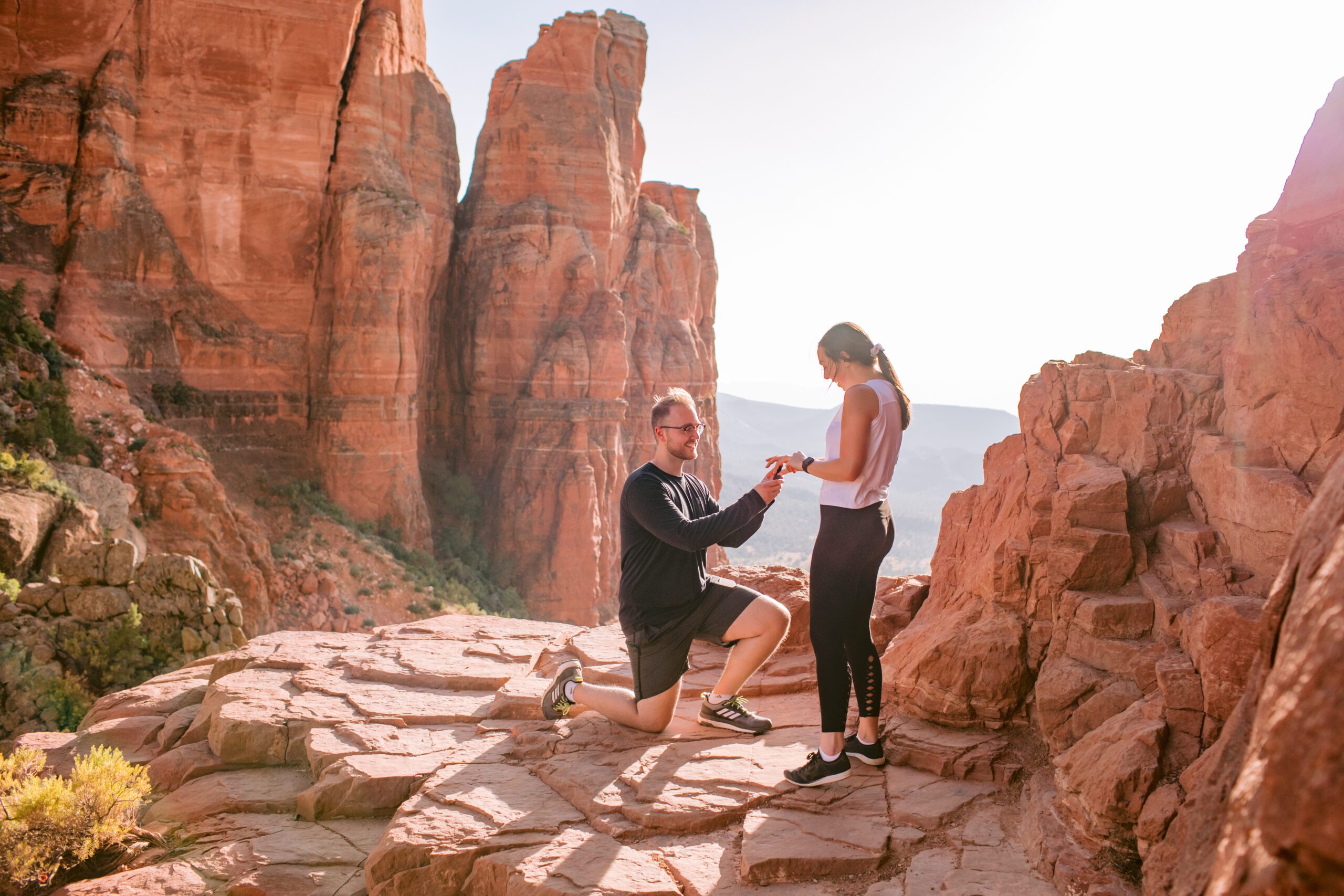 A man is on his knee asking his girlfriend to marry him on the top of Cathedral Rock on a ledge.