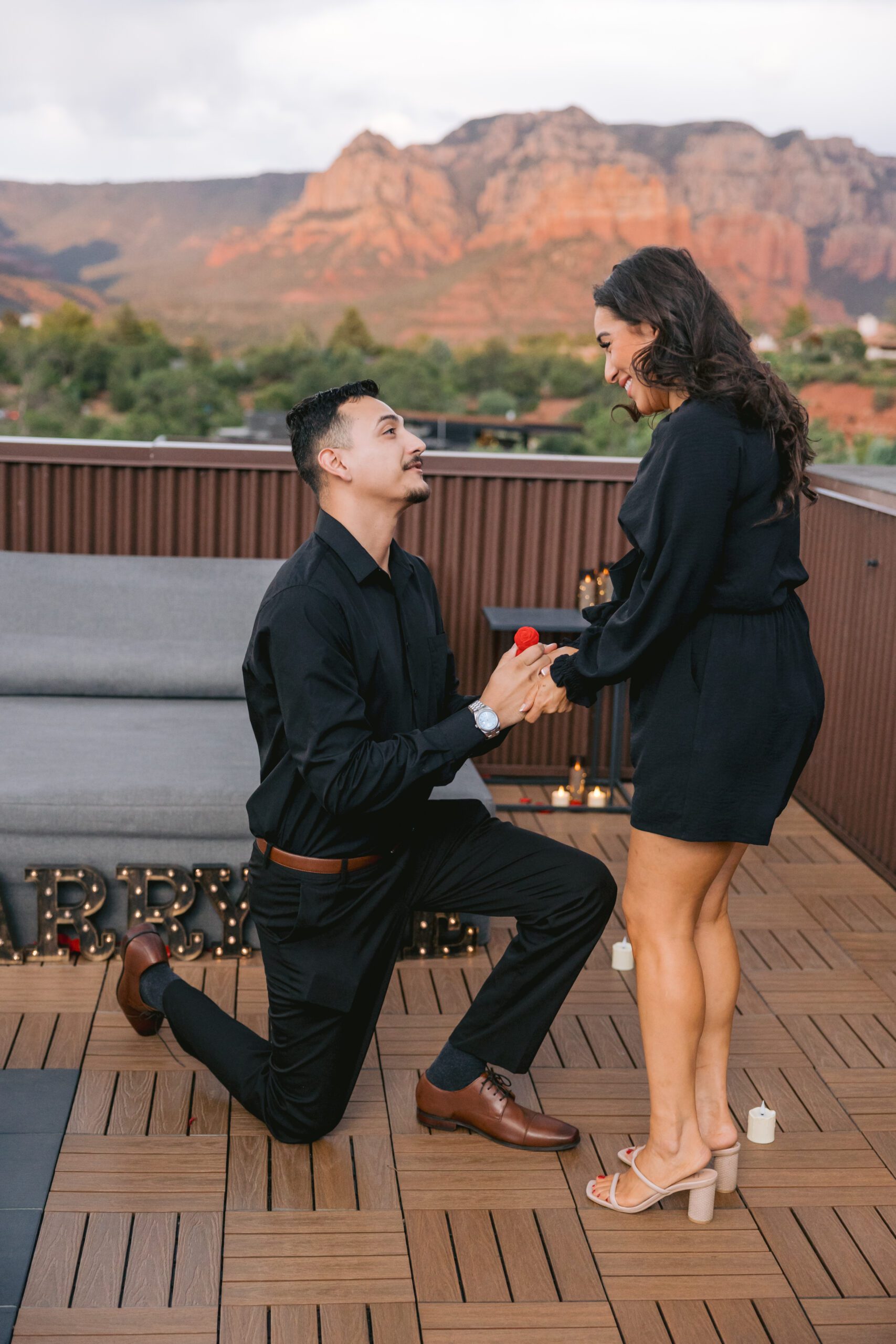 A man in a black shirt and black pants proposes to a woman in a dress on the rooftop of Ambiente Resort.