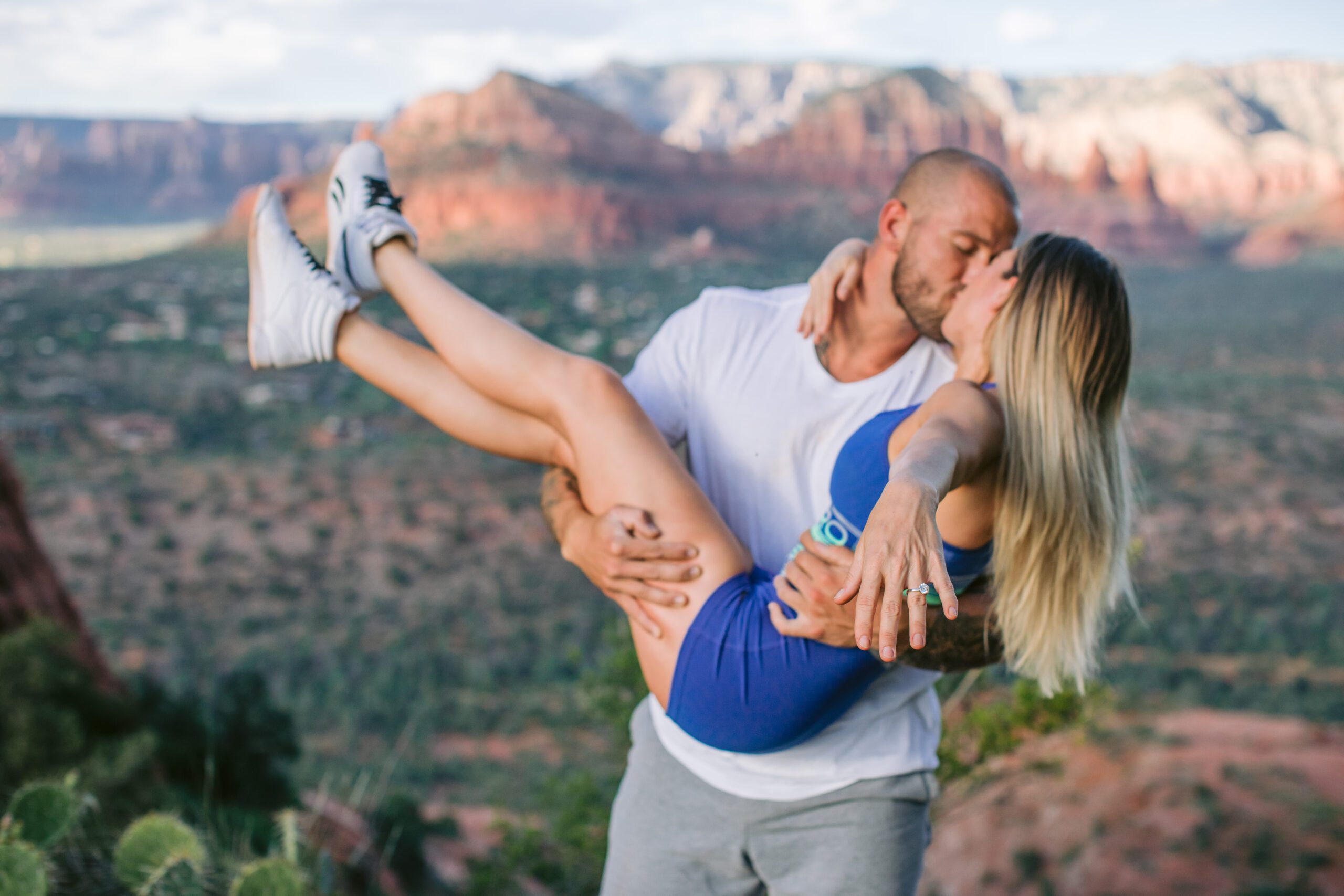 A man holds his girlfriend in his arms and she holds out her hand that has a new engagement ring on it.