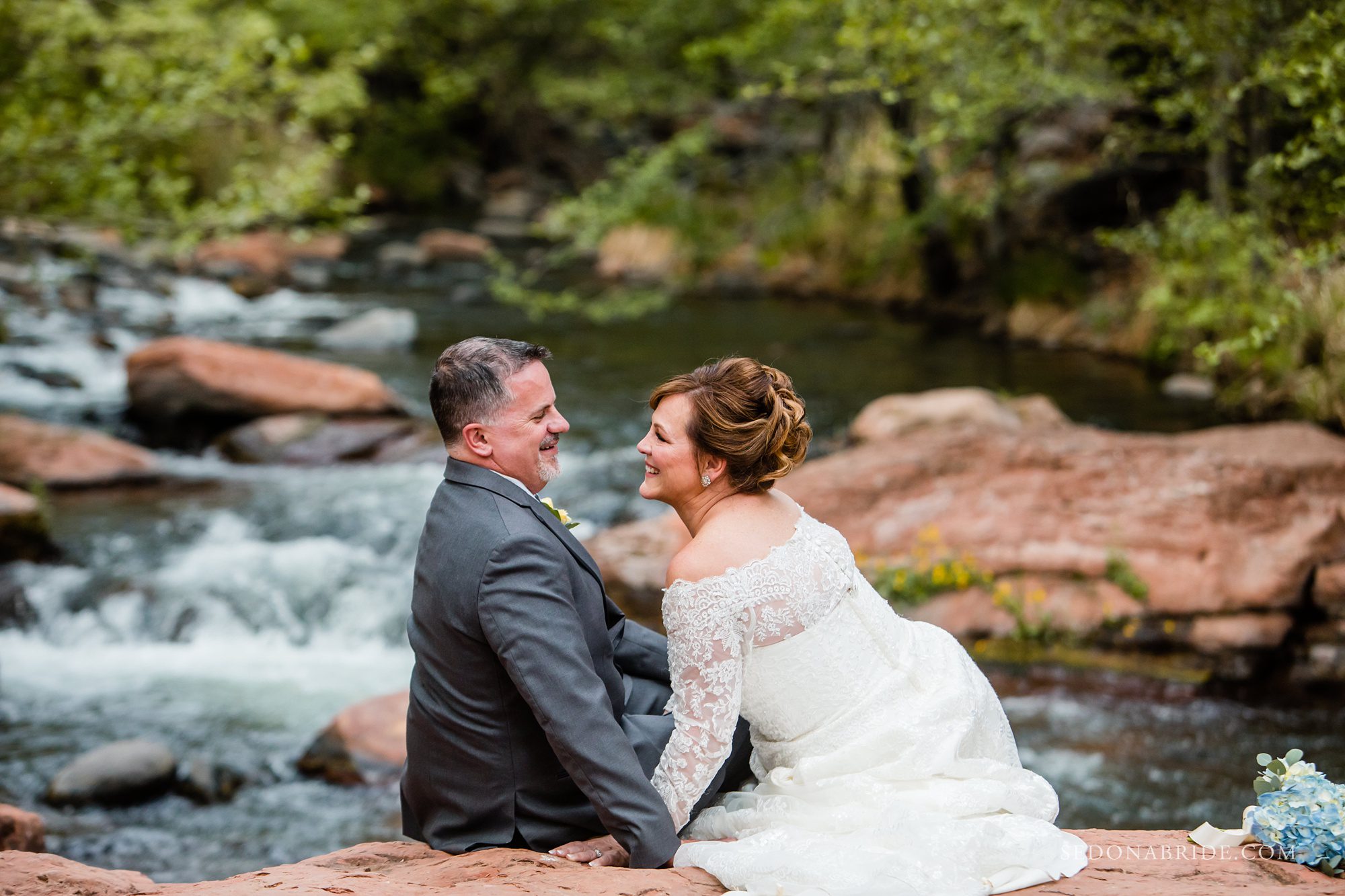 Bride and groom sitting on the red rocks with Oak Creek in the background 