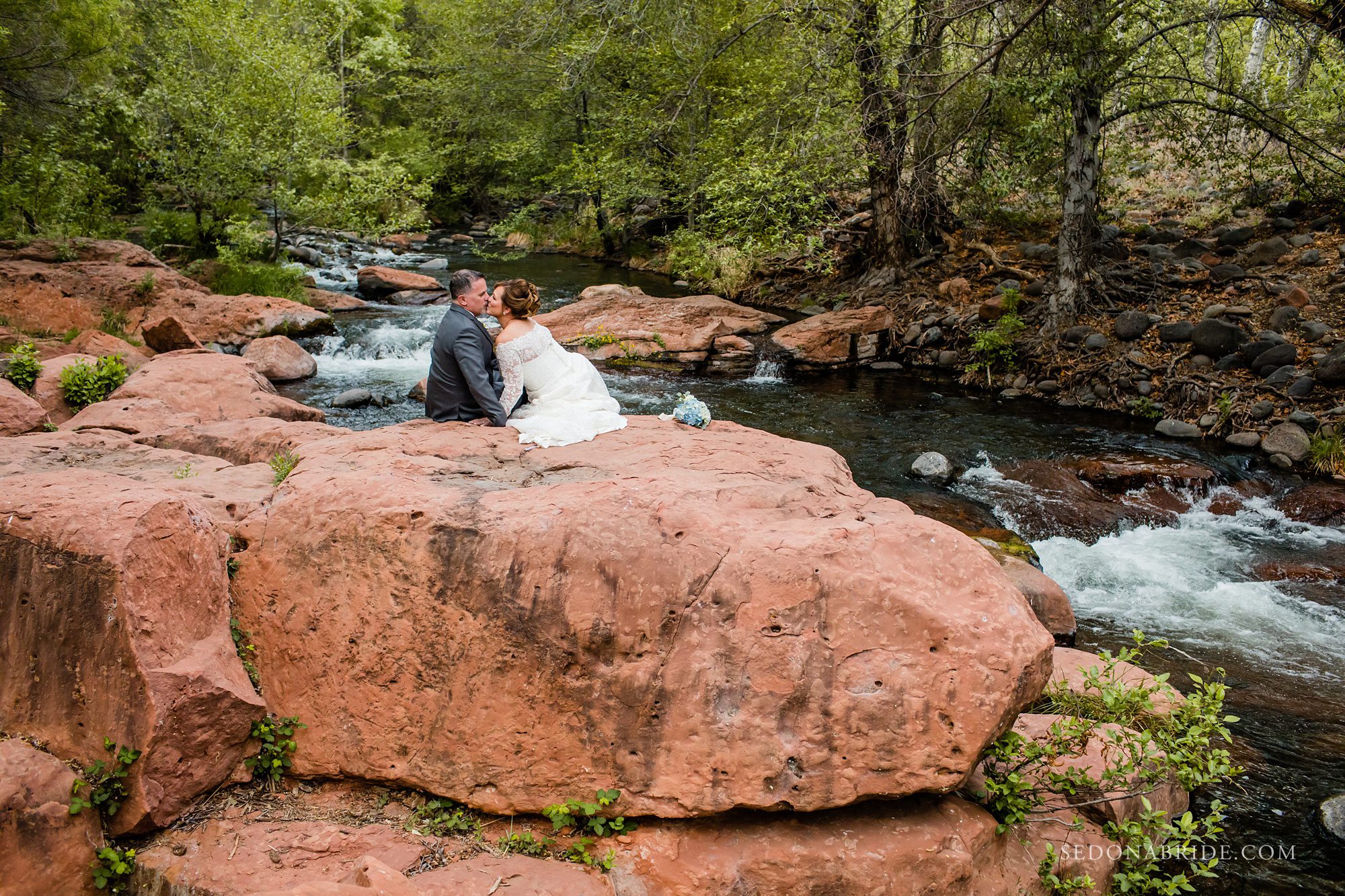 Bride and groom sitting on the red rocks with Oak Creek in the backdrop at Serenity Point at L'Auberge