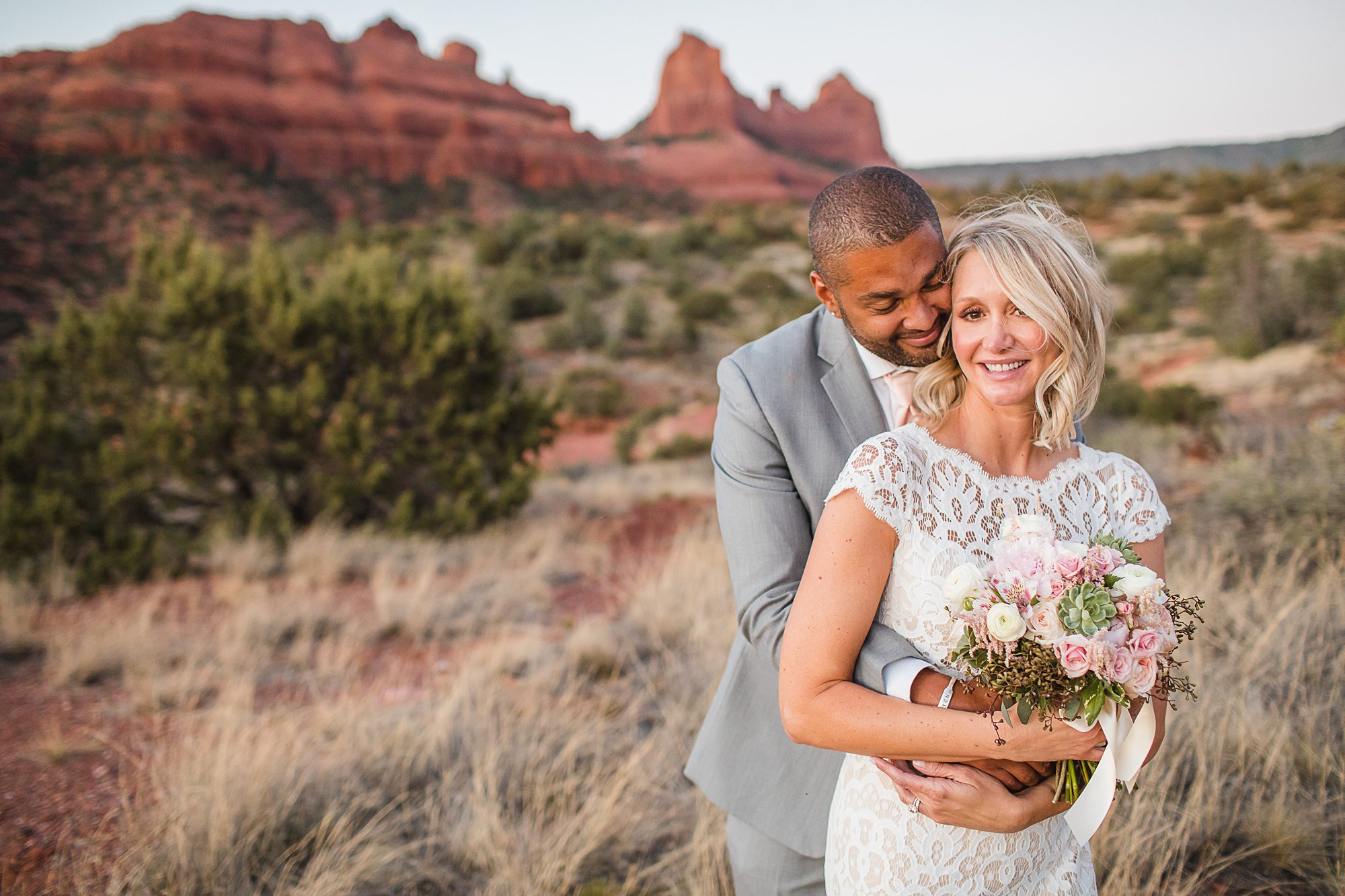 Groom in a grey suite wraps his arms around his bride with the red rocks of Oak Creek in the backdrop 