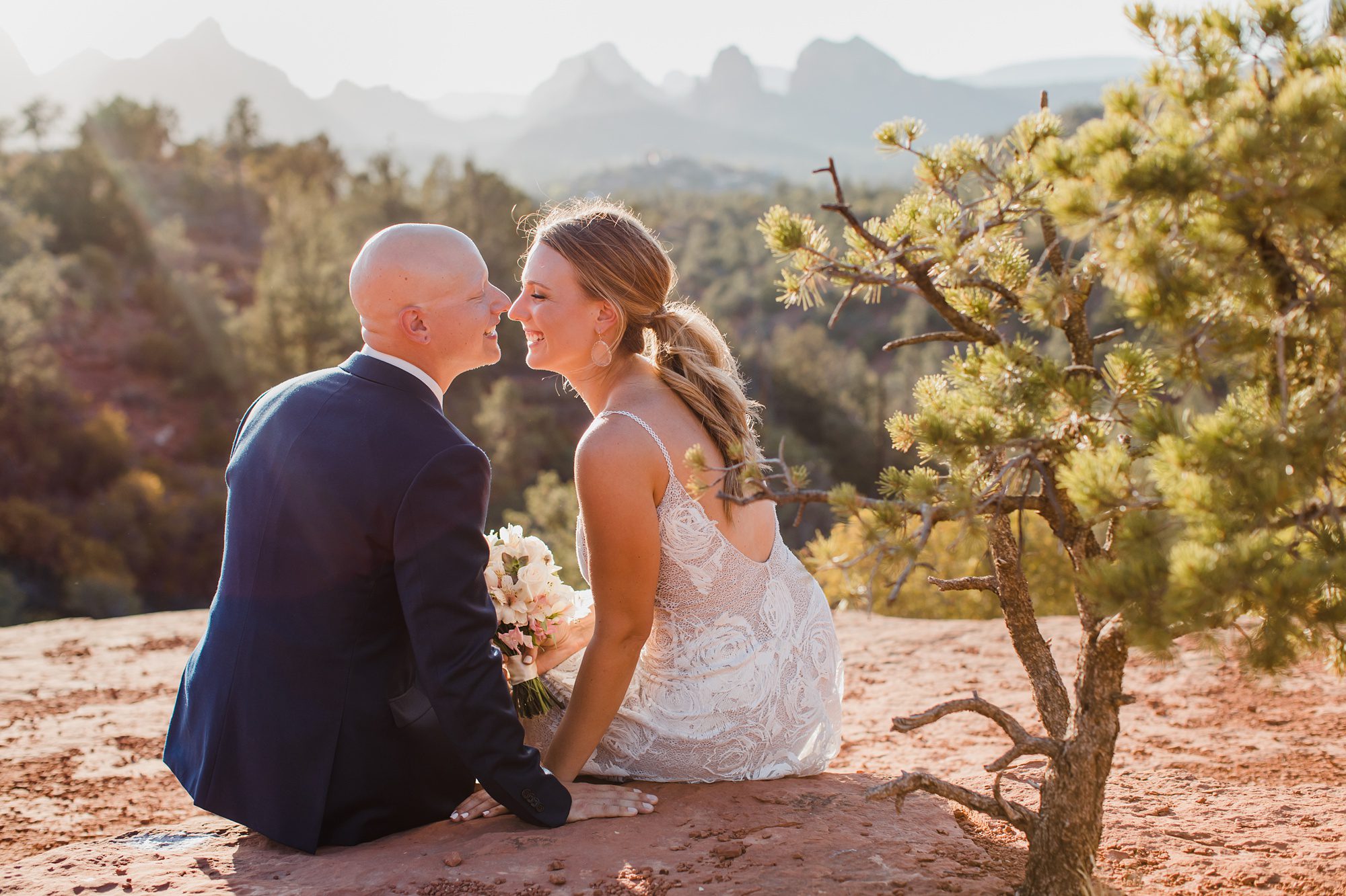 A couple sits on red rocks with uptown Sedona as a backdrop for their wedding photos