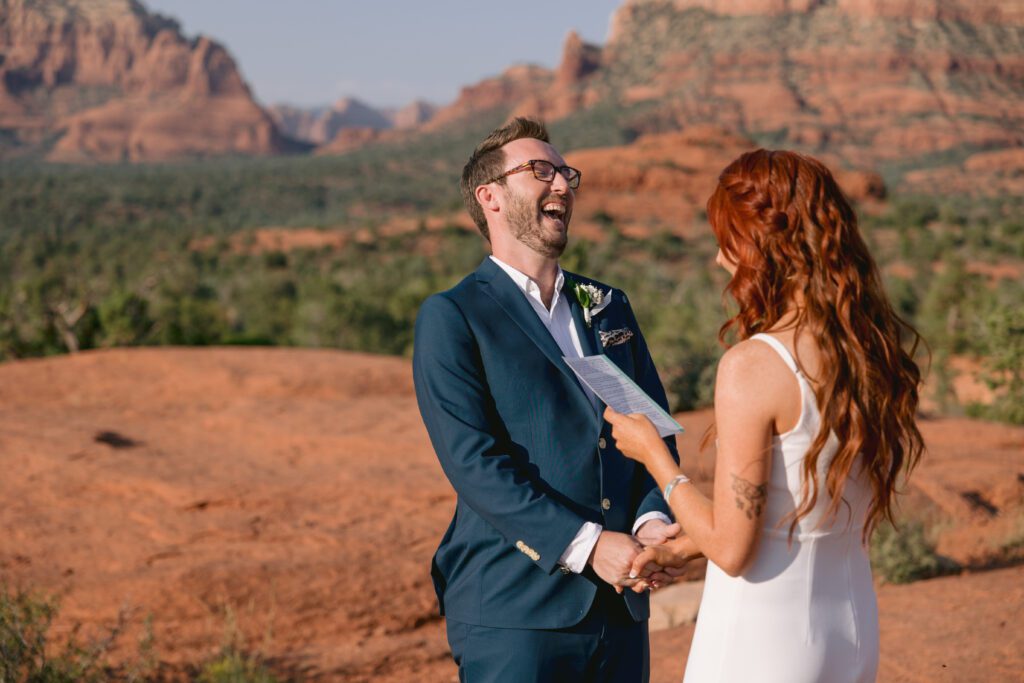 A groom wearing glasses laughs while the bride reads her vows with the red rocks of Sedona in the background during their elopement ceremony at Bell Rock.