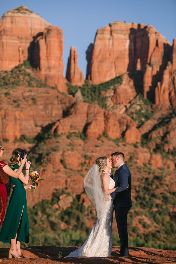 Bride and groom share their first kiss as guests applaud with the glow of Cathedral Rock in the background.