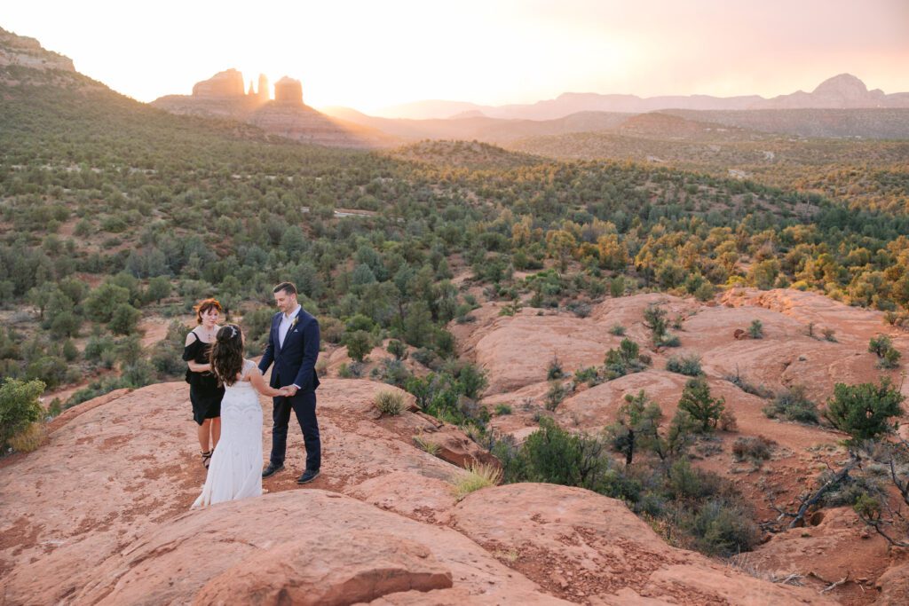 A man wearing a suite holds the hands of his bride wearing a white dress while an officiant performs their wedding ceremony at Bell Rock with Cathedral Rock in the background.