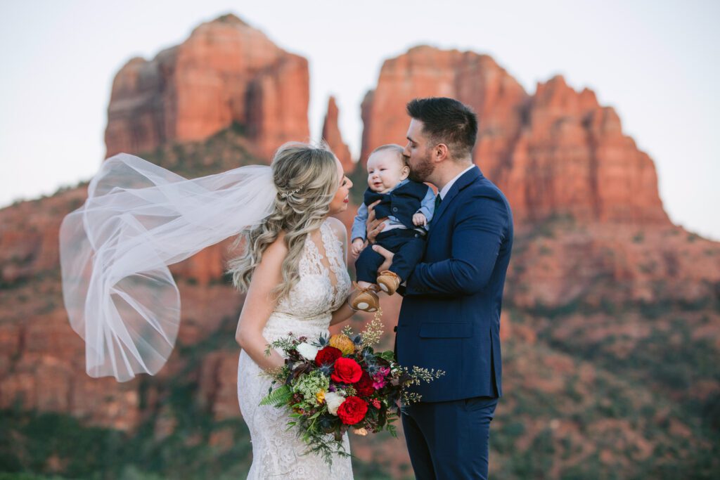 Bride looks at her baby boy who is held by the groom with Cathedral Rock in the backdrop.