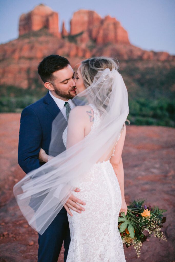 Bride and groom kiss with Cathedral Rock in the backdrop. The bride is wearing a long veil and holding a colorful bouquet.
