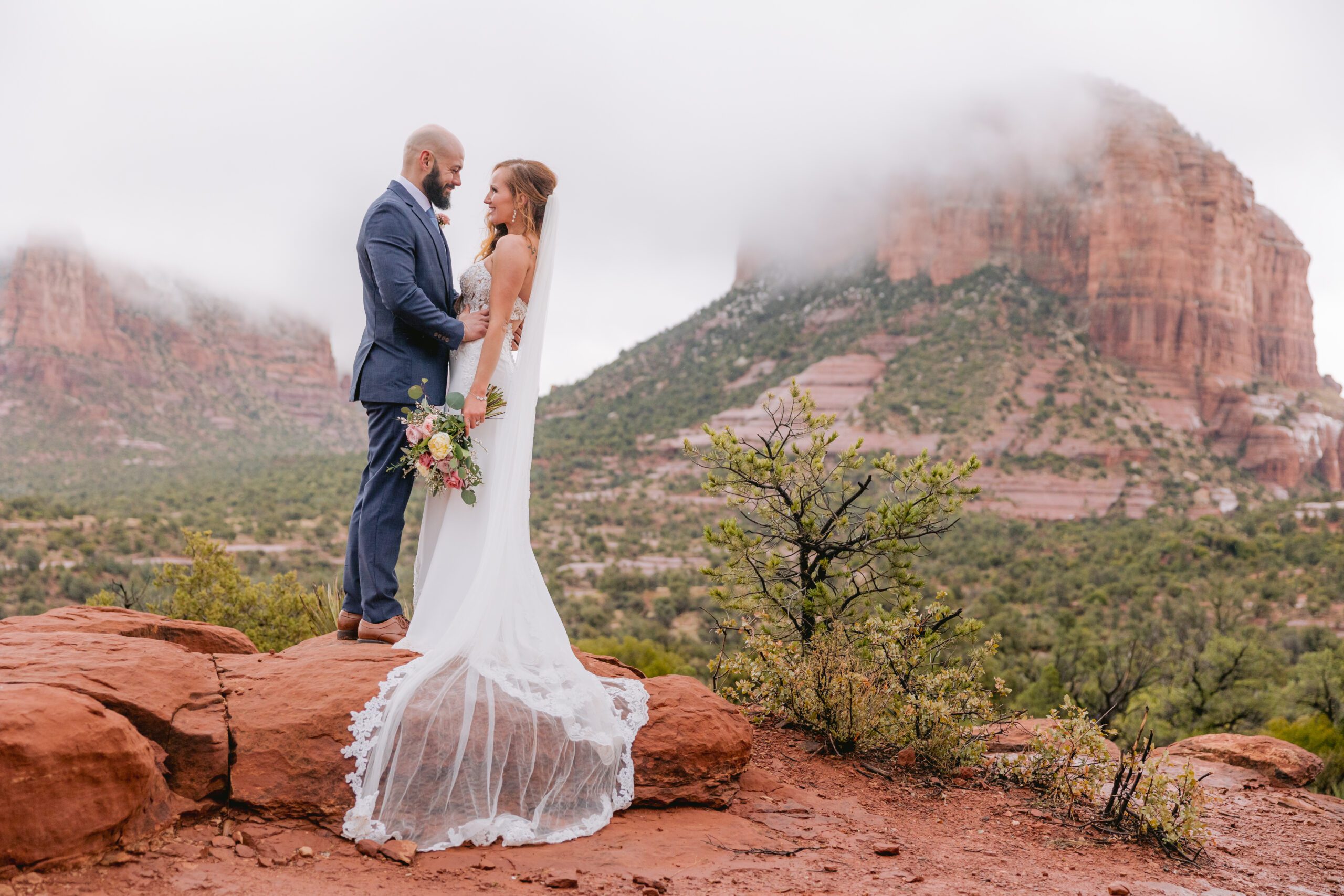 Sedona elopement photos of a bride and groom look at each other and smile following their elopement at Bell Rock. The bride is wearing a long white dress with a long veil that drapes over the red rocks. Behind them are low cloud cover over the red rocks. The bride is holding a floral bouquet with soft pinks and ivory and greens. Her hair is red and the groom has a dark beard.