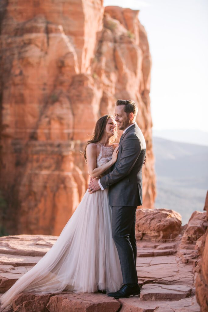 A woman with long brown hair in a wedding dress smiles at her a man dressed in a grey suit smiling off into the distance. They are holding each other and the red rocks of Cathedral Rock are behind them along with the valley during their elopement in Sedona.