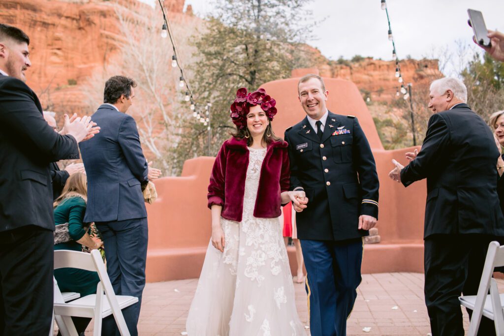 Micro-wedding with a bride wearing a red floral crown, red jacket and white wedding dress and her groom in a military suit walking down their wedding aisle with guests clapping during there elopement at Enchantment Resort. 
