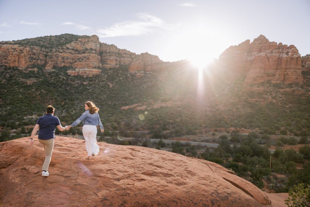 Sun sets on the ridge of the red rocks while a couple walks holding hands for their Sedona engagement photos.