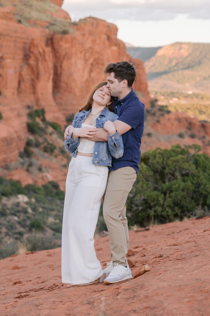A couple embraces. The woman with brown hair has her back to a man with brown hair's chest. She's wearing a denim jacket and white top and white pants. The man is wearing a blue shortsleeved shirt and khaki pants.