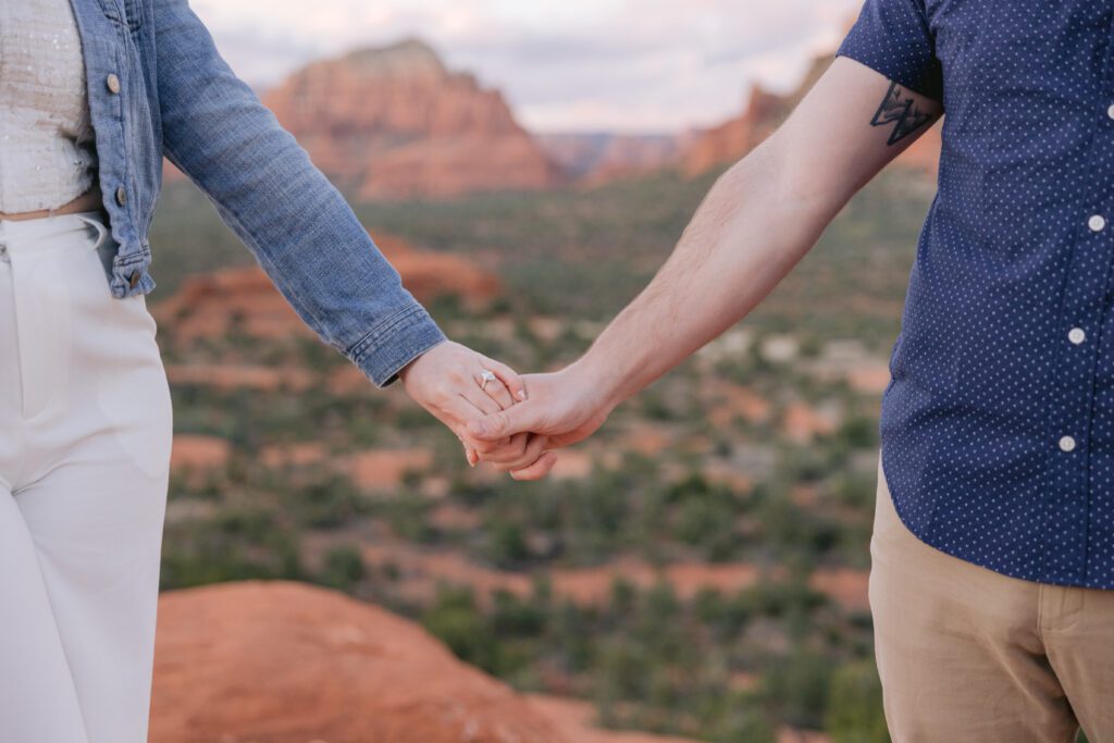 A couple holds hands with the engagement ring showing and the Sedona red rocks behind the couple.