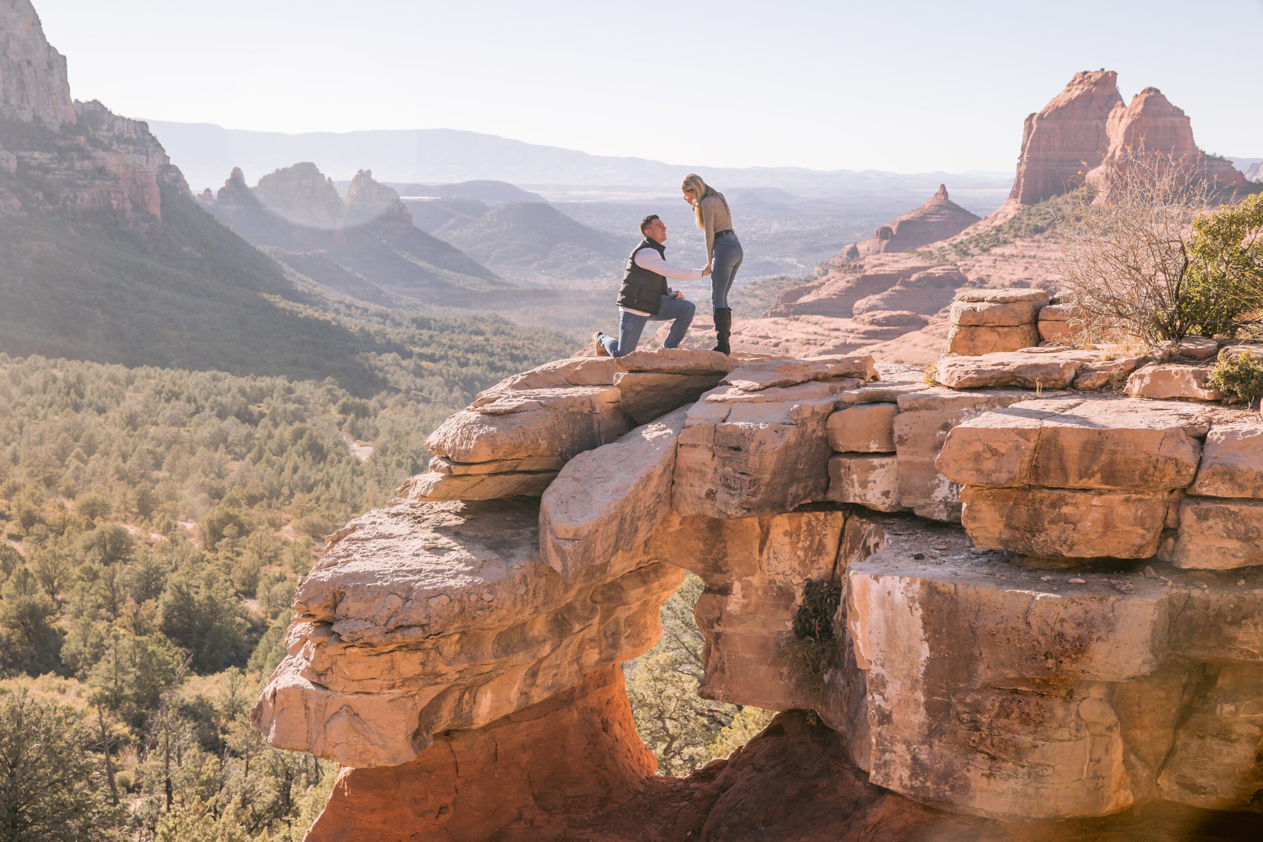 A man is down on one knee to propose to a woman standing next to him on a famous ledge at Merry Go Round Rock in Sedona. There is a hole in the rock below them and a valley with red rocks that surround them.
