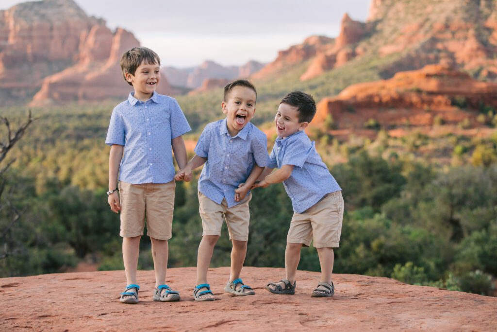 Young brothers wearing blue shirts and tan shorts hold hands and laugh with the red rocks behind them for their family photos.
