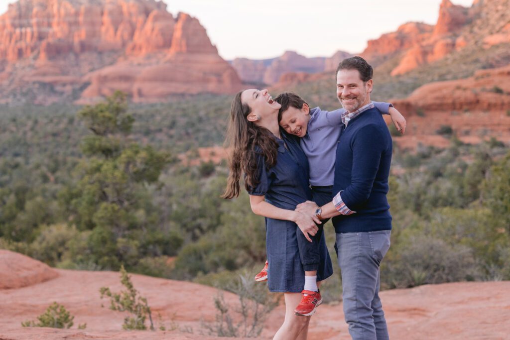 Mom laughs while holding her son and her son is laughing as well and the dad is looking at the camera.
