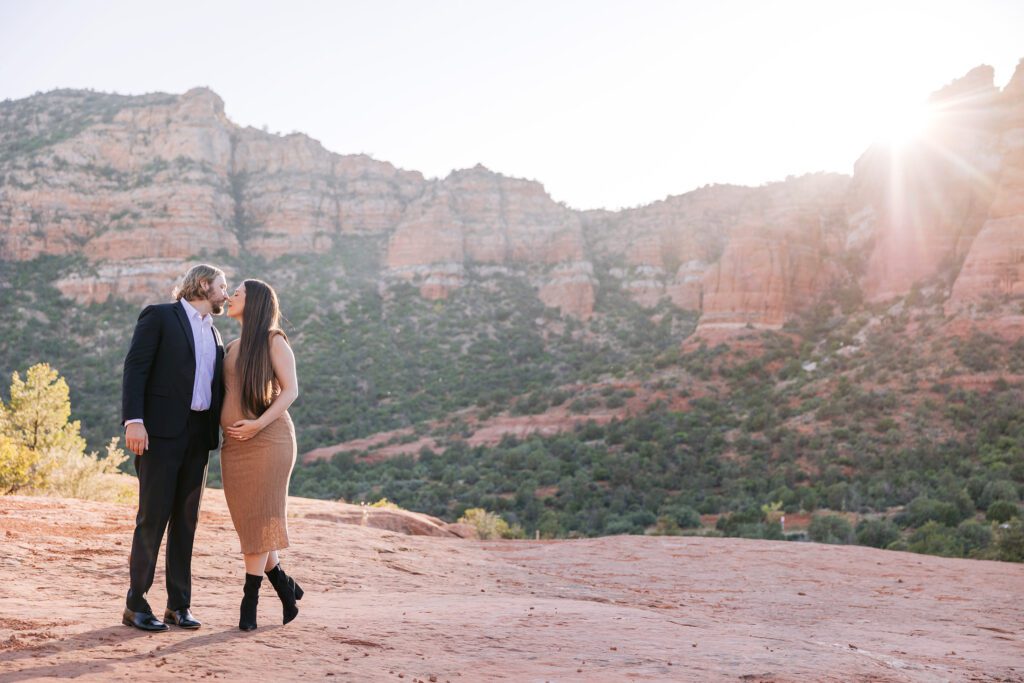 A man and a pregnant woman kiss with the setting sun behind them on the red rocks.