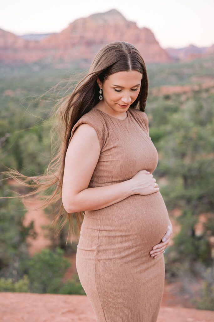 A portrait of a pregnant woman with long brown hair, wearing a tan dress and holding her belly for a maternity photos in Sedona.