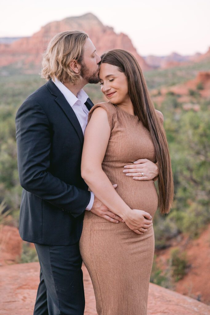 A man with shoulder length hair kisses his wife while holding her belly for maternity photos in Sedona.