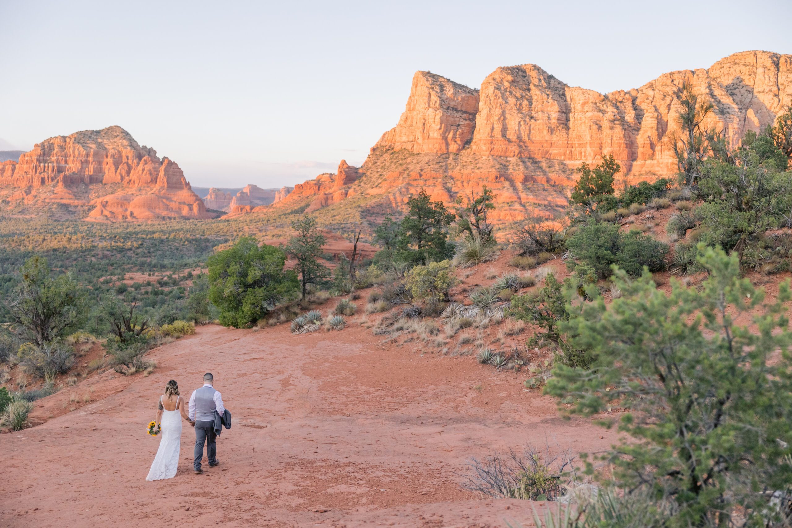 Bride walks on the red rocks with her groom holding his jacket on his arm. The red rocks glow in the background with the warmth of the sunset after their Bell Rock wedding.